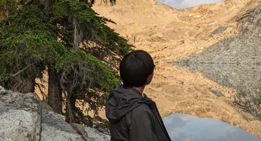 A person faces away from the camera and looks out at an alpine lake, reflecting the mountains surrounding it. 